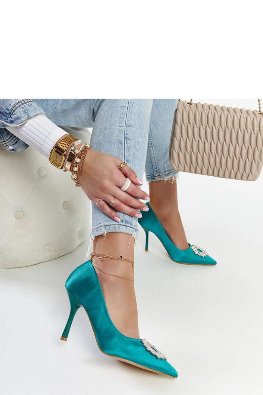 Stylish woman wearing green stiletto heels with satin finish and zirconia, paired with jeans and a chic handbag.