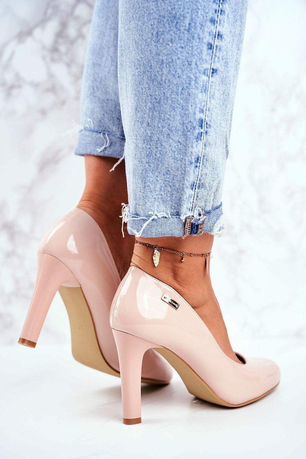 Blush pink high heels with a shiny finish and ankle strap worn with cuffed jeans, showcasing classic style and elegance.