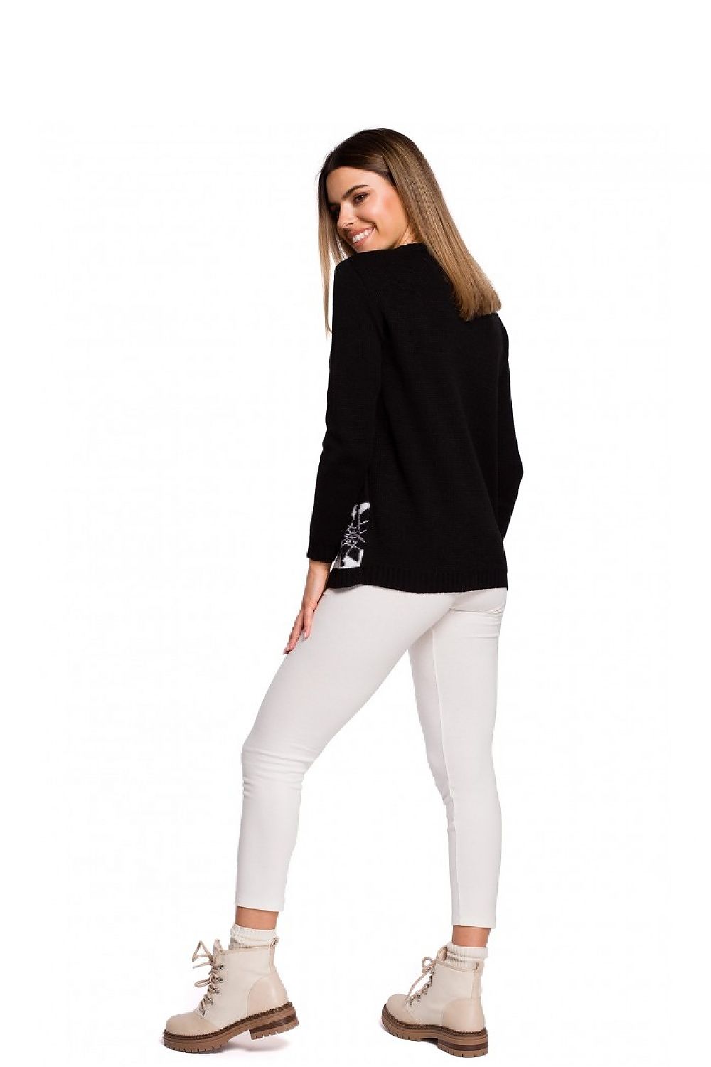 Woman in black pullover with reindeer motif, white pants, and beige boots, smiling and looking over shoulder, showcasing winter fashion.