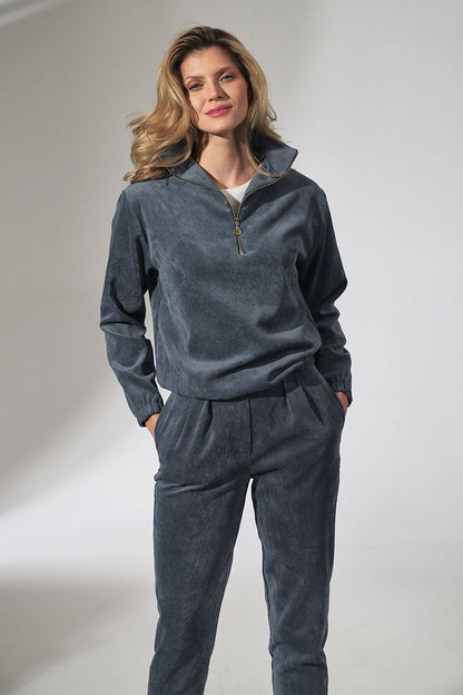 Woman wearing gray velour tracksuit with zippered top and high collar, hands in pockets, styled with loose curls and confident smile.