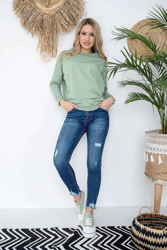 Woman wearing a stylish green pullover and jeans, standing in a chic, modern room with plants and boho decor, casual fashion look.