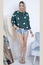 Model wearing a green heart-patterned pullover and denim shorts, reflecting casual style and comfort in a modern living room setting.