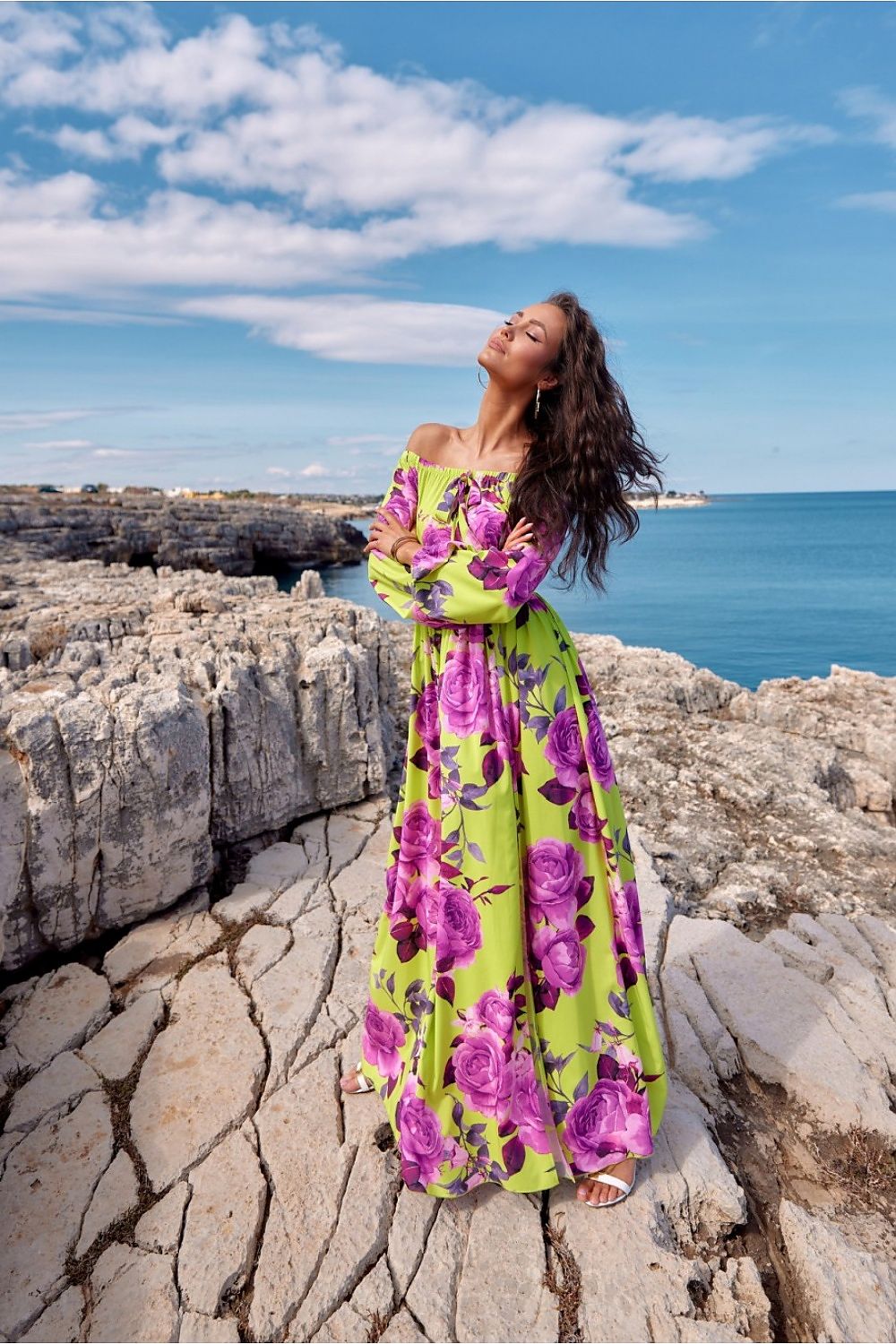 Woman in vibrant floral long dress by the sea on rocky coast, with open shoulders and tied neck detail, embracing the view.