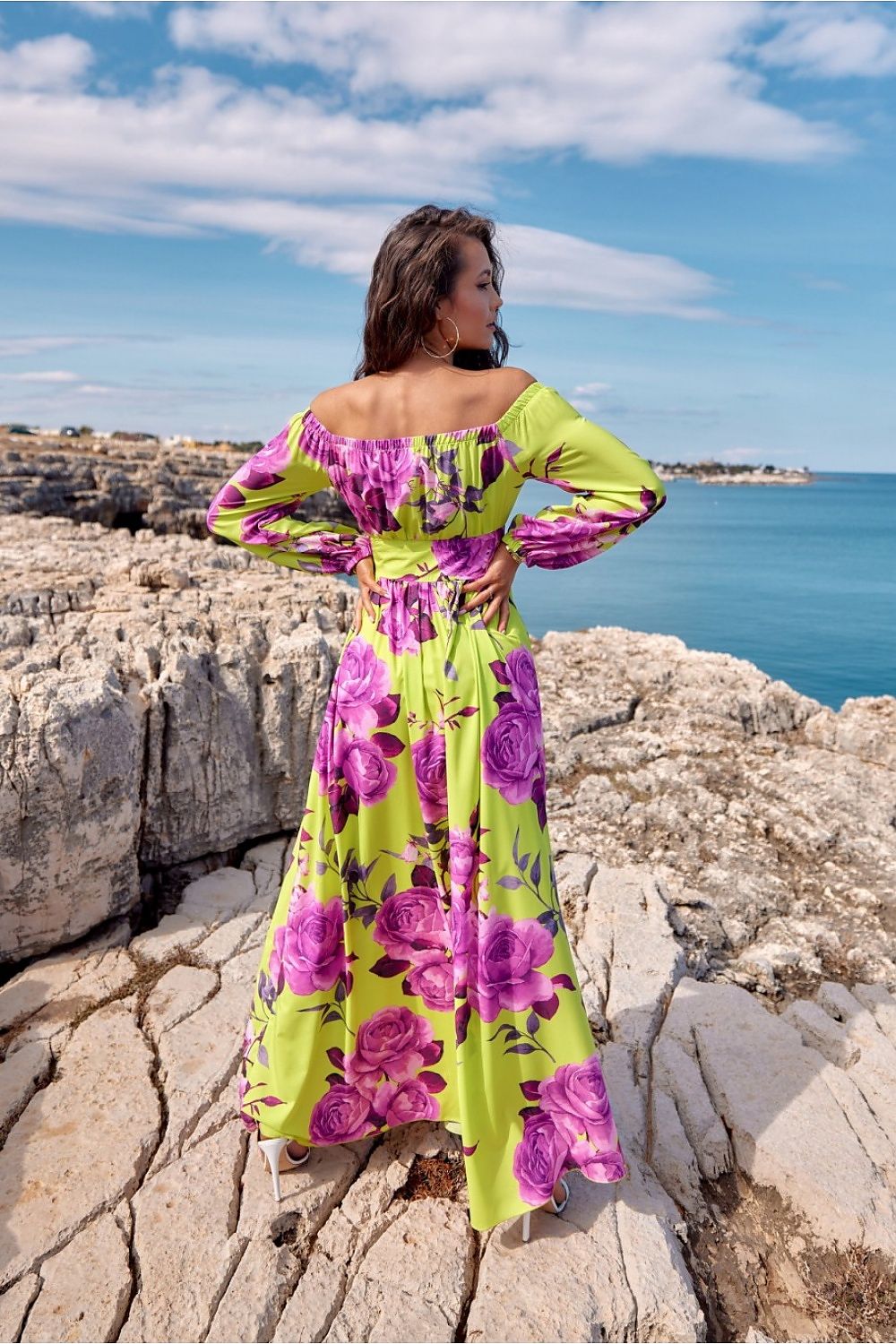 Model wearing off-shoulder floral maxi dress with waist tie and leg slit by the sea, showcasing vibrant colors and elegant design.