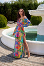 Vibrant floral maxi dress with open shoulders and side slit, featuring a decorative neck tie and elastic shoulder straps, model by a fountain.