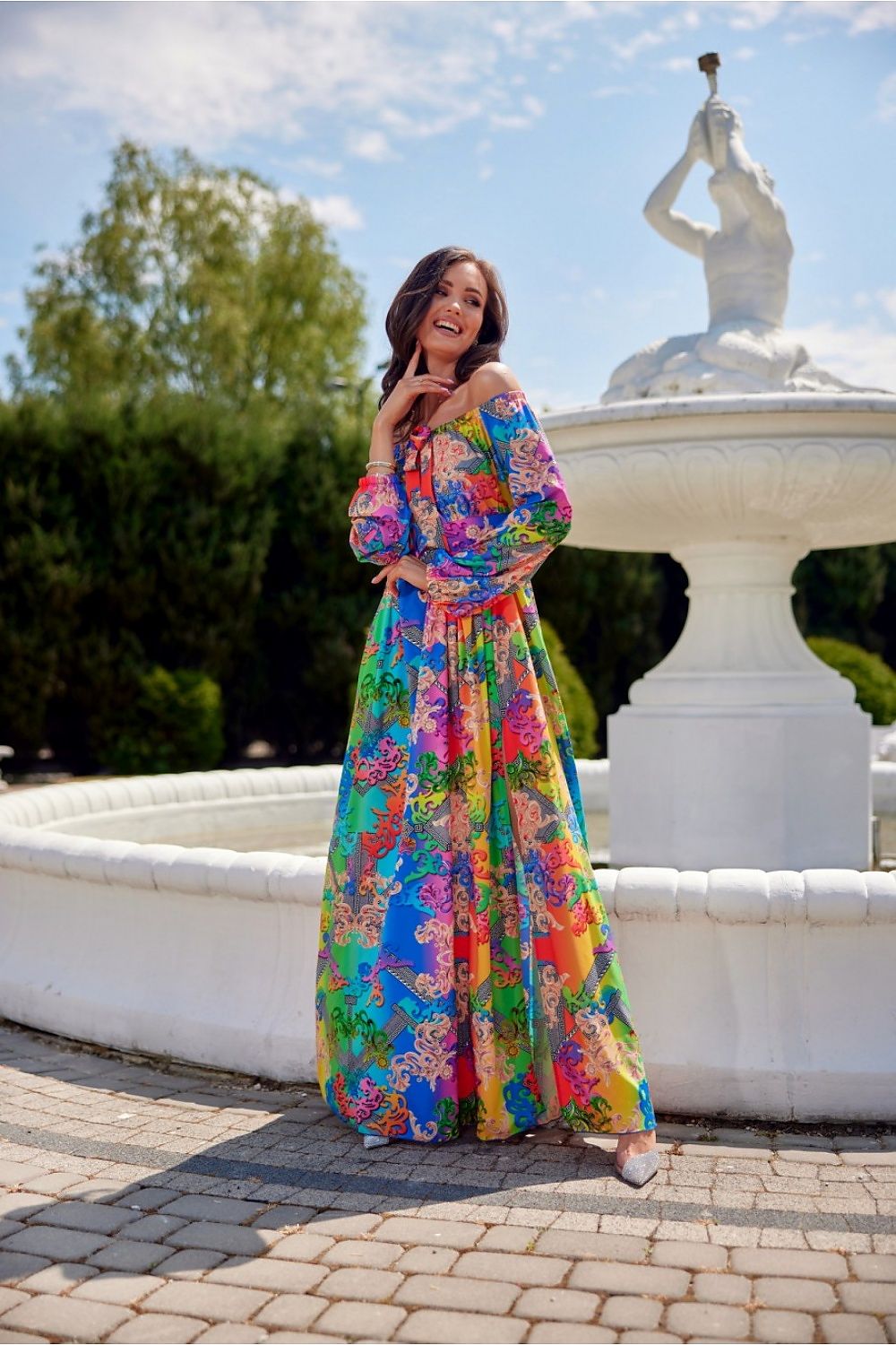 Colorful Roco Fashion maxi dress with open shoulders, standing by a fountain, elegant and vibrant design.